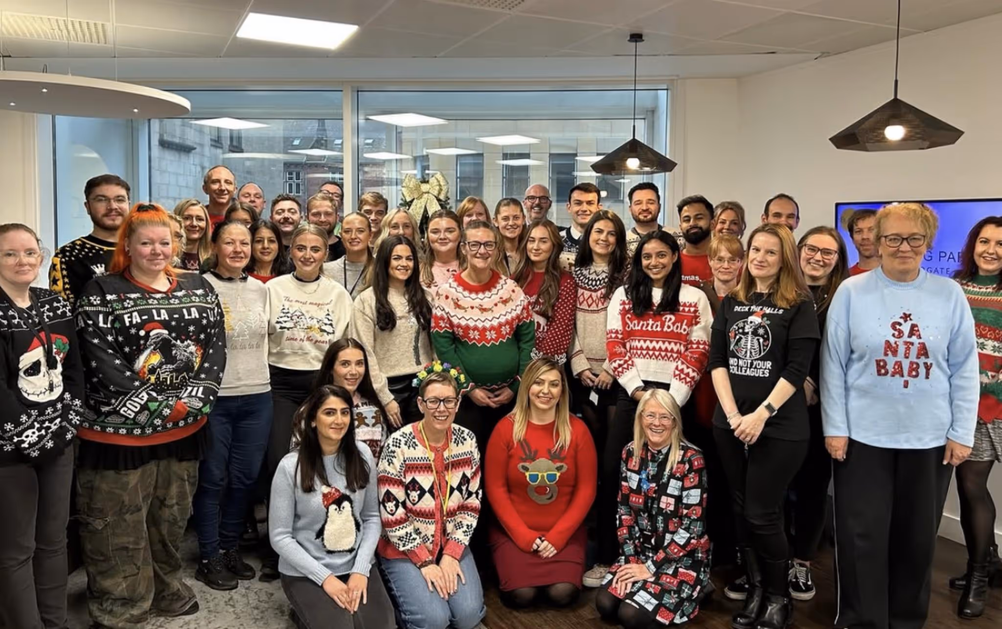 Group of people in colorful Christmas sweaters posing indoors with holiday decorations behind them.