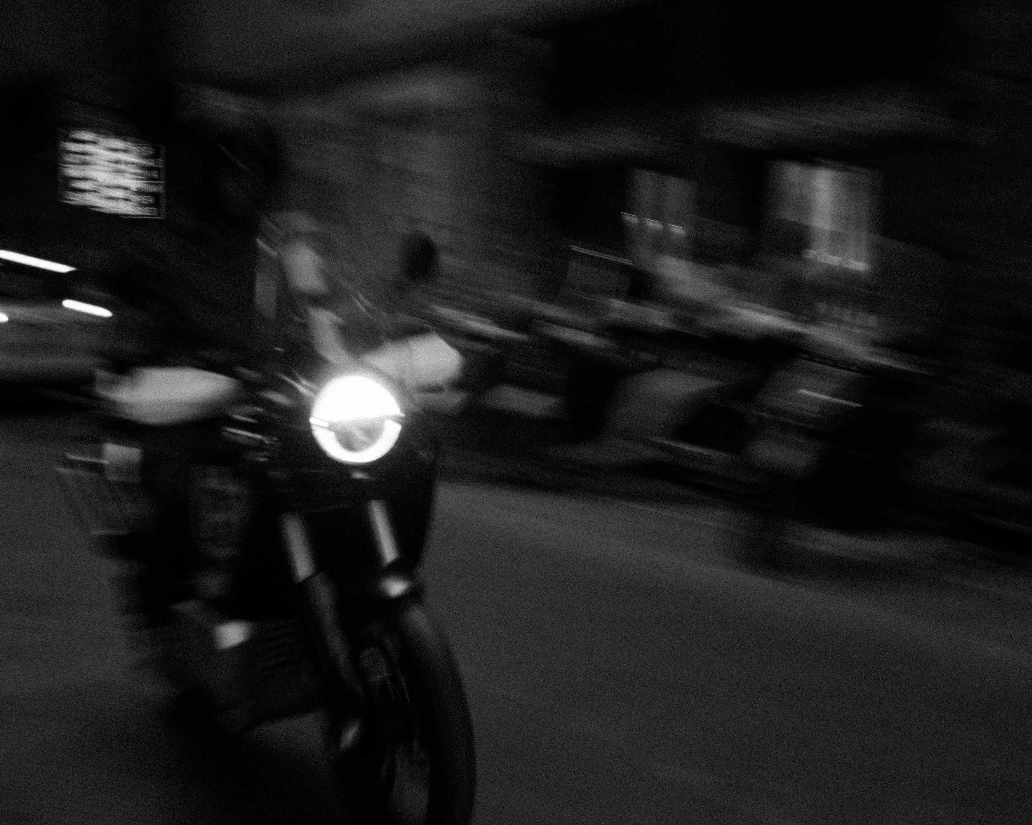 Blurry black and white image of a motorcycle with its headlight on, moving in a dimly lit urban environment.