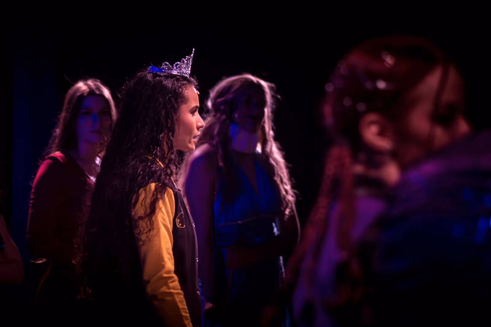 Young woman with curly hair wearing a tiara surrounded by other people in dim, colorful stage lighting.