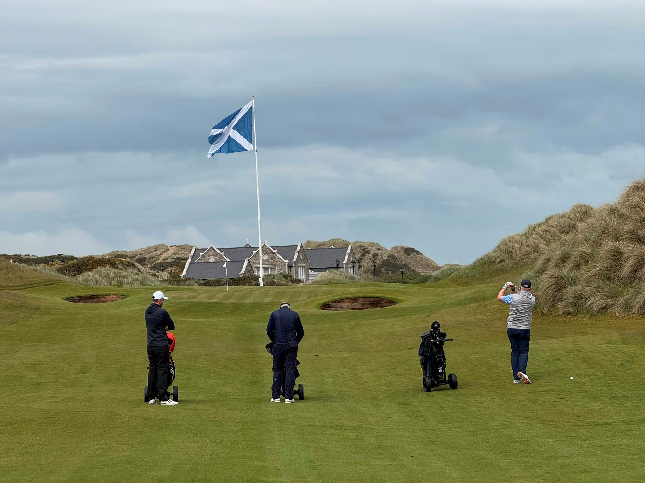 Four golfers on a green fairway near sand bunkers with a Scottish flag and clubhouse in the background.