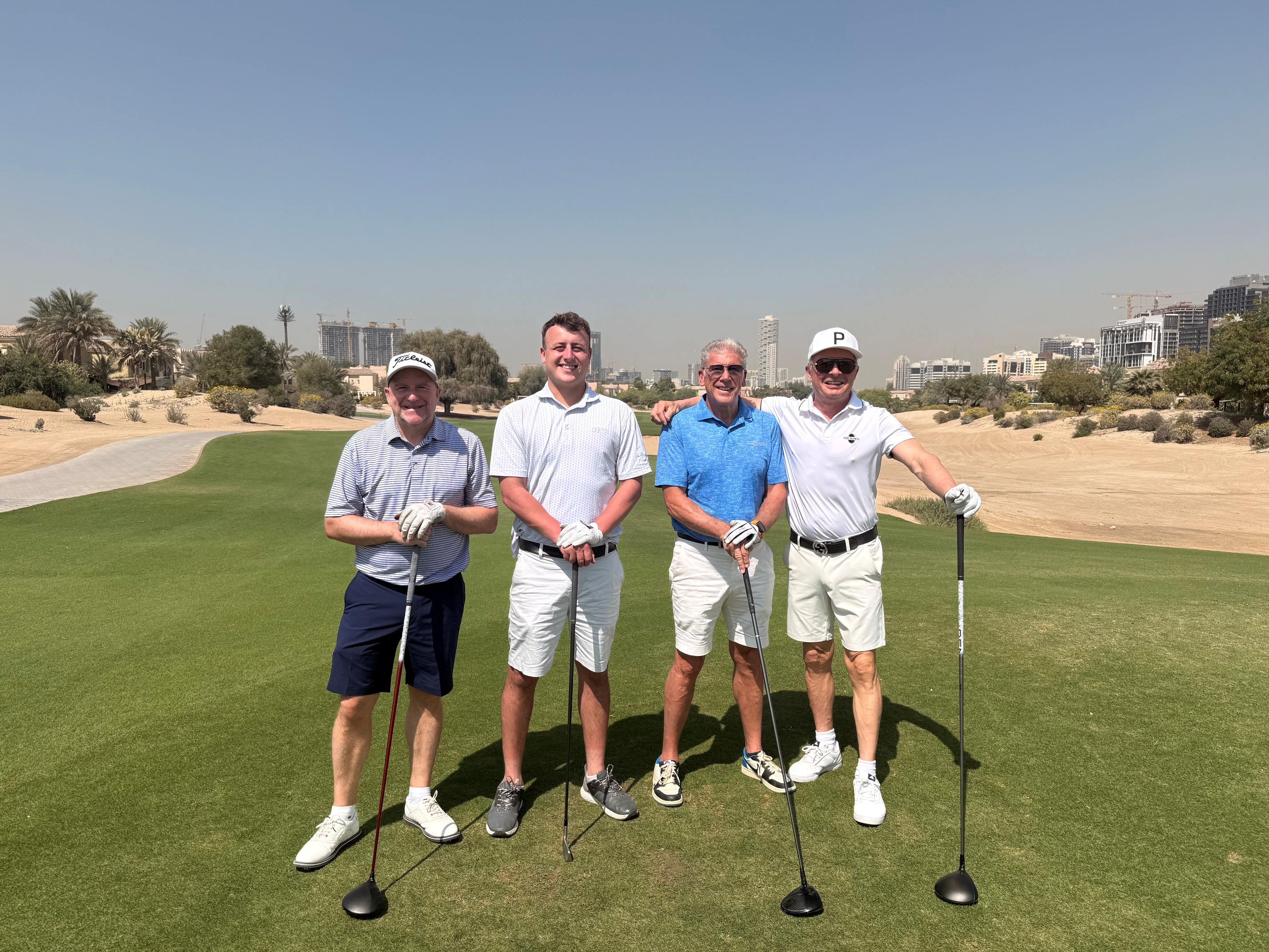 Four men standing on a golf course holding clubs, dressed in collared shirts and shorts, smiling at camera.