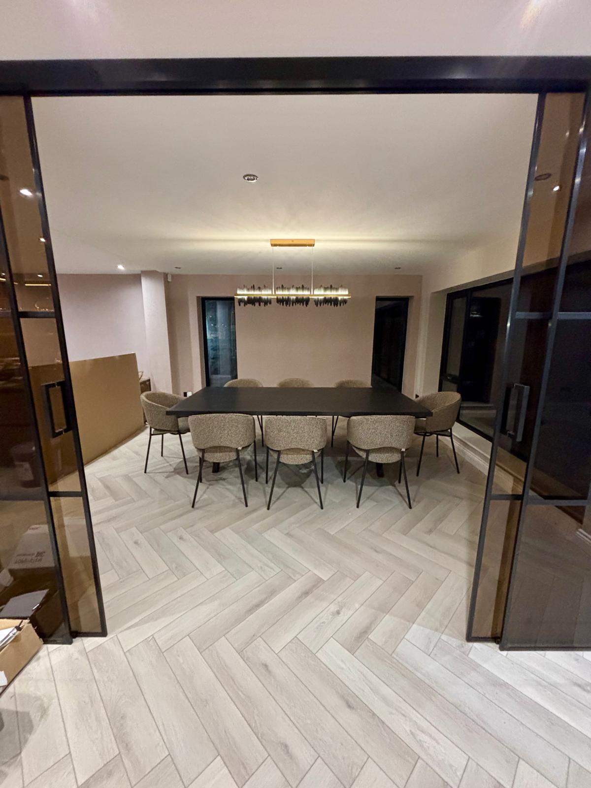 Modern dining room with a black rectangular table surrounded by eight beige upholstered chairs and herringbone light wood flooring.
