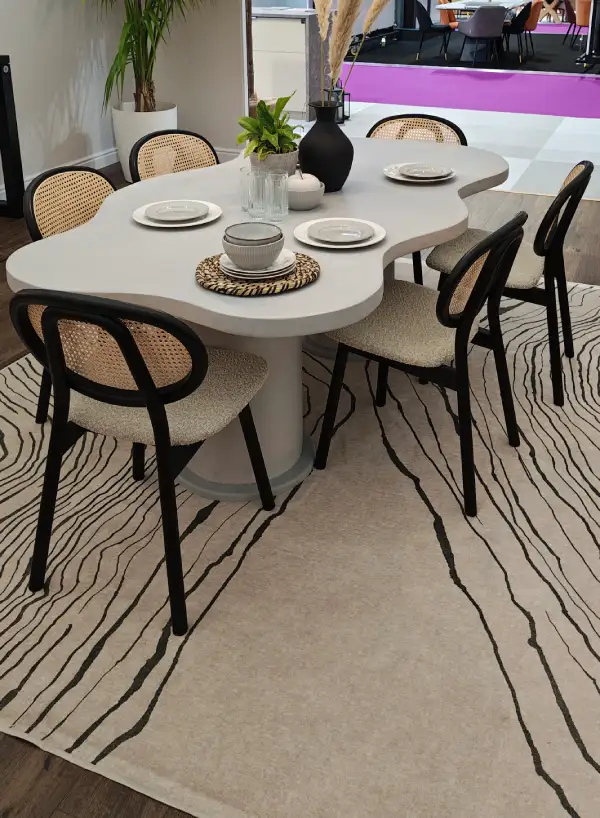 Modern white dining table with irregular shape, set with plates and bowls, surrounded by six chairs with black frames and beige woven backs on a beige rug with black abstract lines.