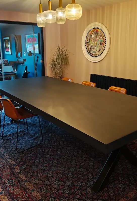 Modern conference room with large dark polished concrete table, leather chairs, patterned rug, hanging glass pendant lights, and artwork on beige walls.