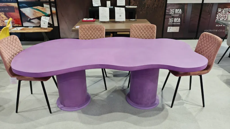 Purple kidney-shaped table with two cylindrical legs surrounded by four pink quilted chairs with black legs in a showroom.