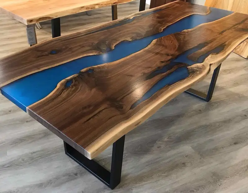 Wooden table with natural edges featuring a blue resin river embedded in the center and black metal legs on a light wood floor.