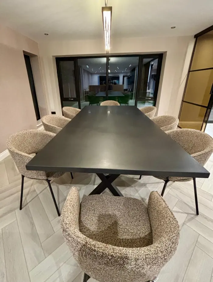 Modern dining room with a long black rectangular table on an X-shaped base surrounded by seven upholstered beige armchairs on light wood herringbone floor.