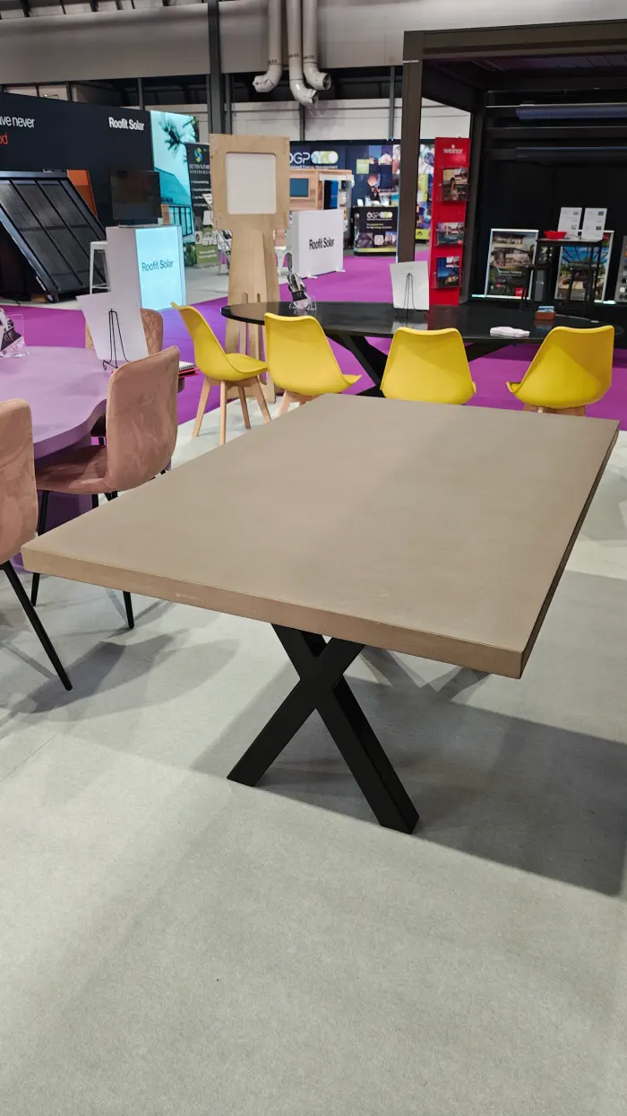 Modern beige rectangular table with black X-shaped legs in a showroom with yellow and pink chairs in the background.