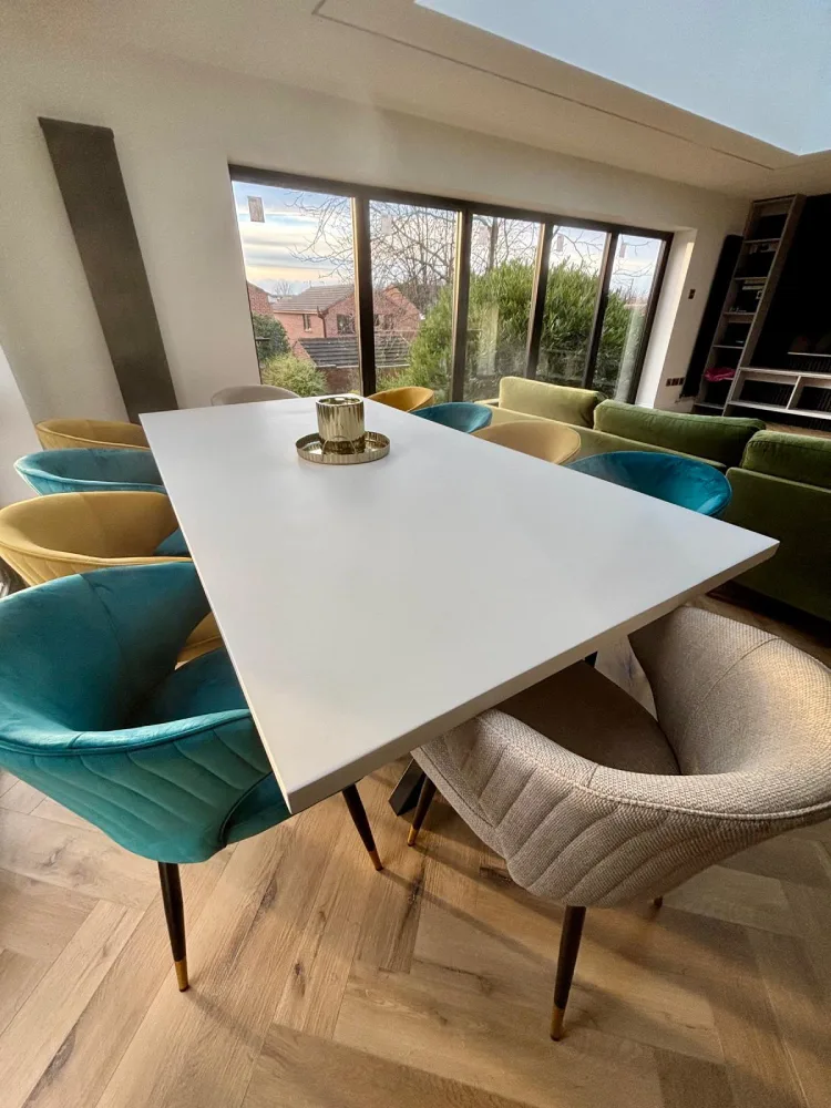 Modern dining table with a white surface surrounded by colorful upholstered chairs in a bright room with large windows.
