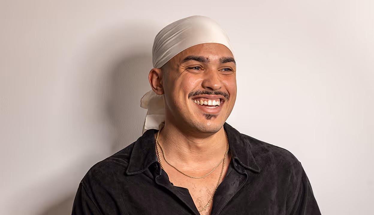 Person in black shirt and white durag smiling brightly against wall