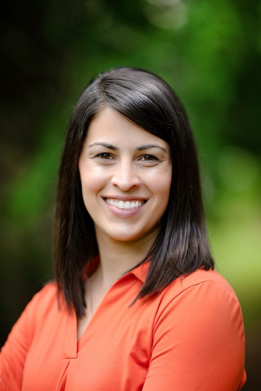 Smiling woman with shoulder-length dark hair wearing an orange top standing outdoors with a green blurred background.