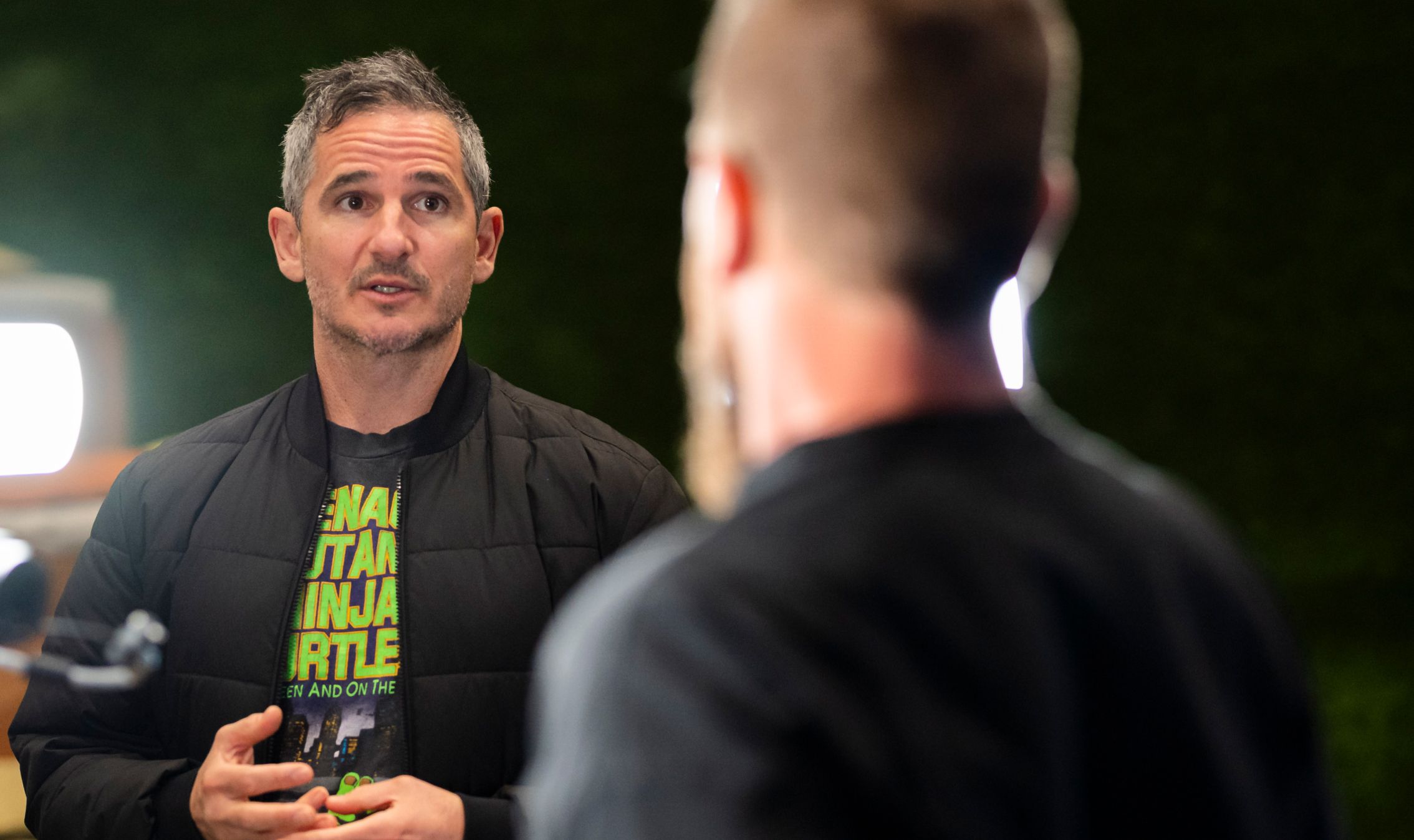 Man with gray hair in a black jacket speaking to another person with a blurred back facing the camera.