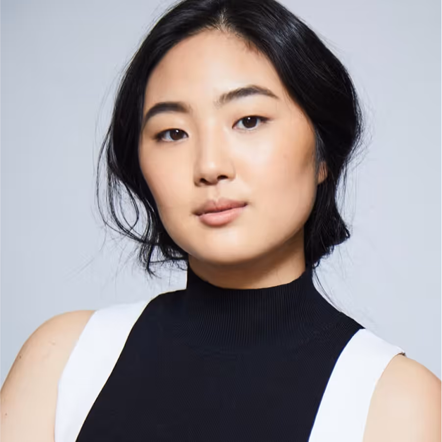 Portrait of a woman with black hair tied back, wearing a sleeveless black and white top, against a plain light gray background.