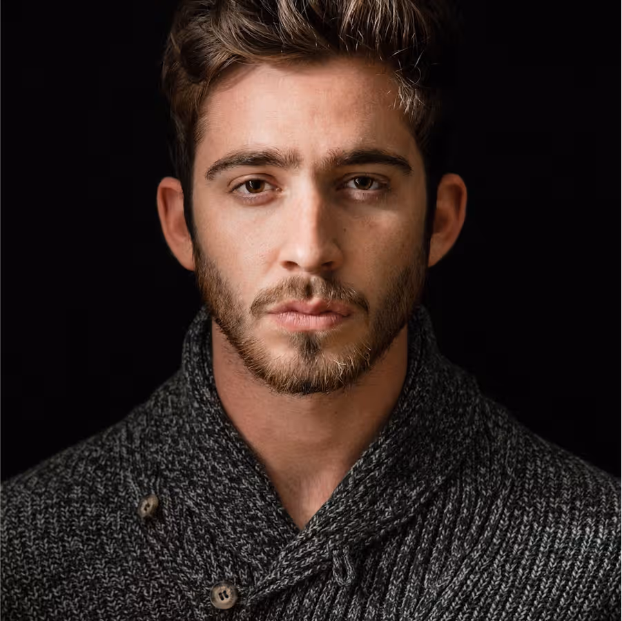 Close-up portrait of a young man with short brown hair, beard, and mustache wearing a dark gray knit sweater against a black background.