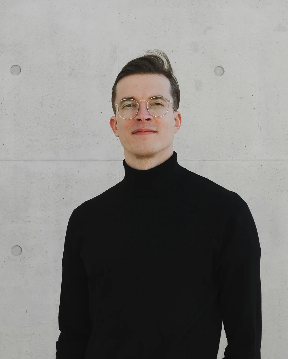 Young man with glasses wearing a black turtleneck sweater standing against a light gray concrete wall.