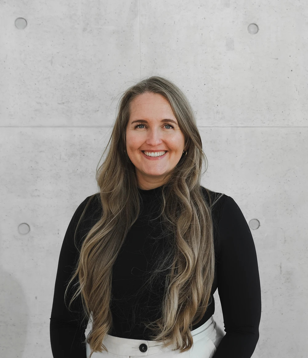 Smiling woman with long wavy blonde hair, wearing a black long-sleeve top and white pants, standing against a gray concrete wall.