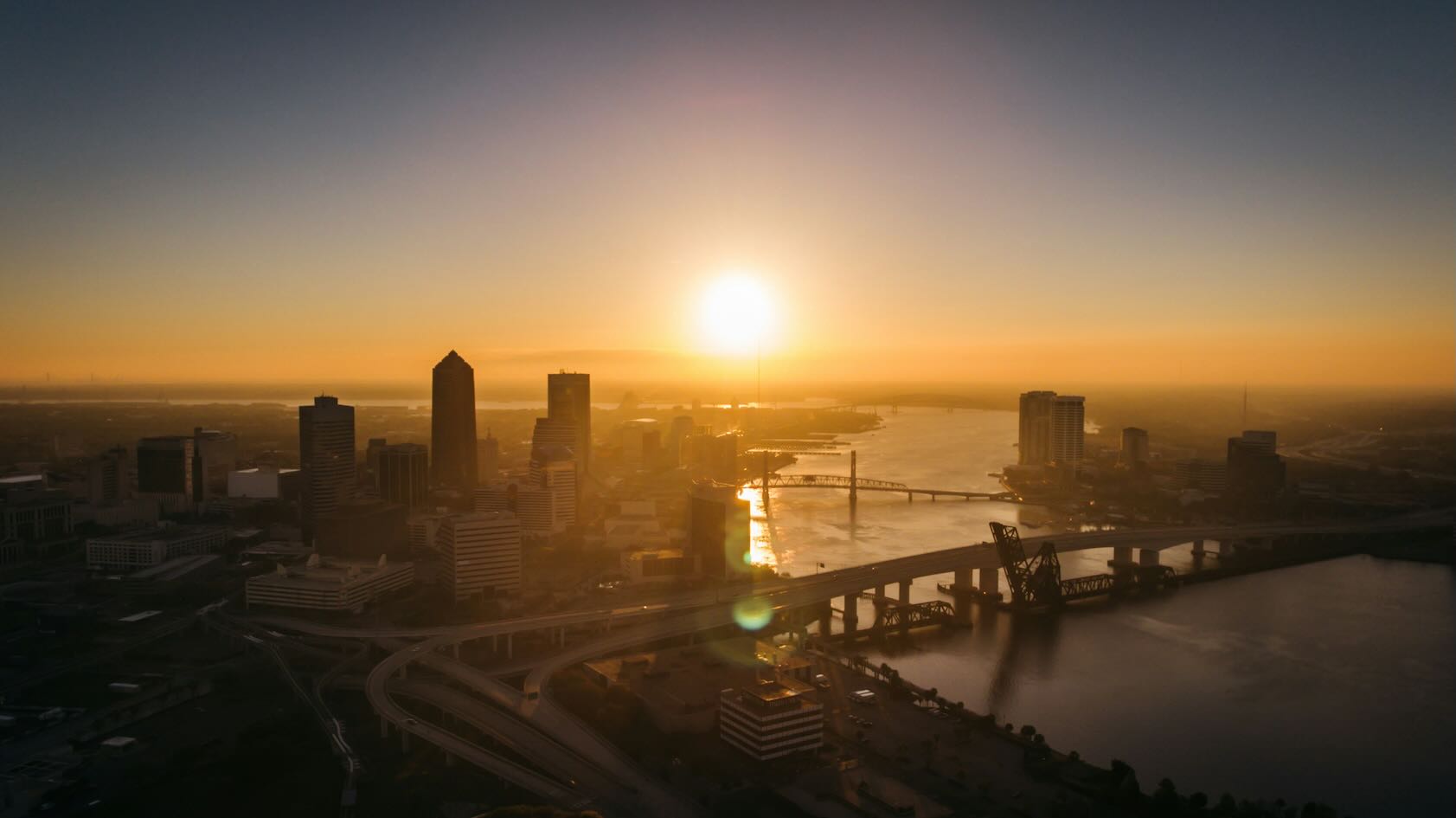 Aerial sunrise over downtown Jacksonville with the St. Johns River and multiple bridges glowing in golden light.