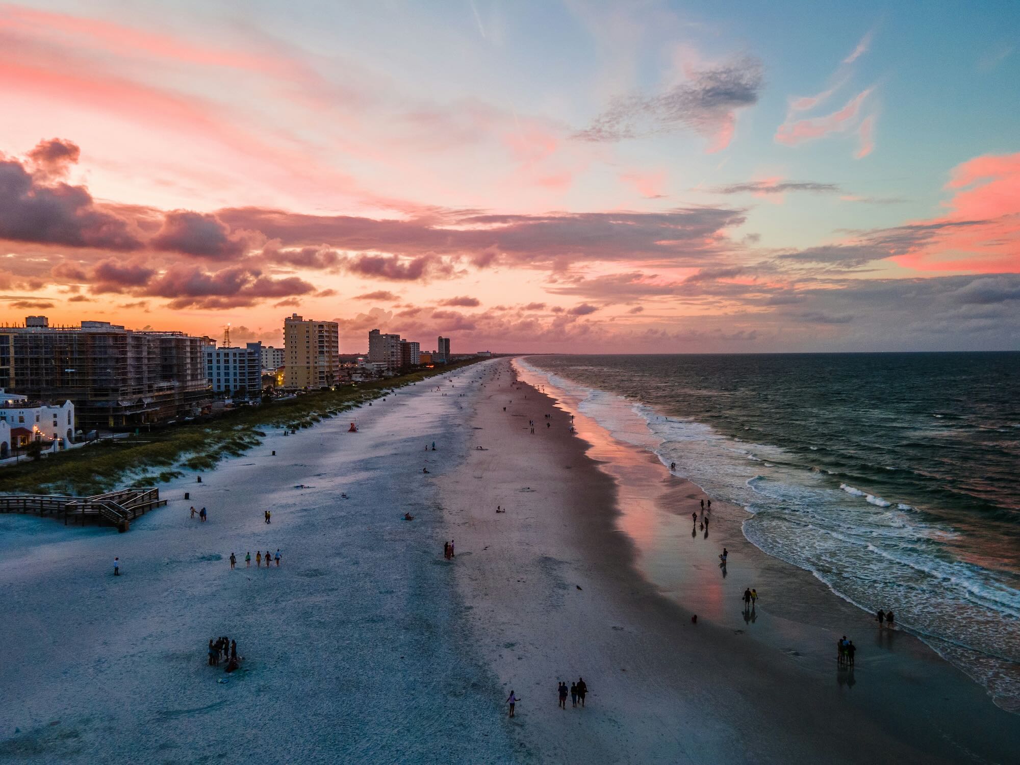 Aerial sunset view of Jacksonville Beach with pastel clouds, condos lining the left, and people walking along the wet shoreline as waves roll in.