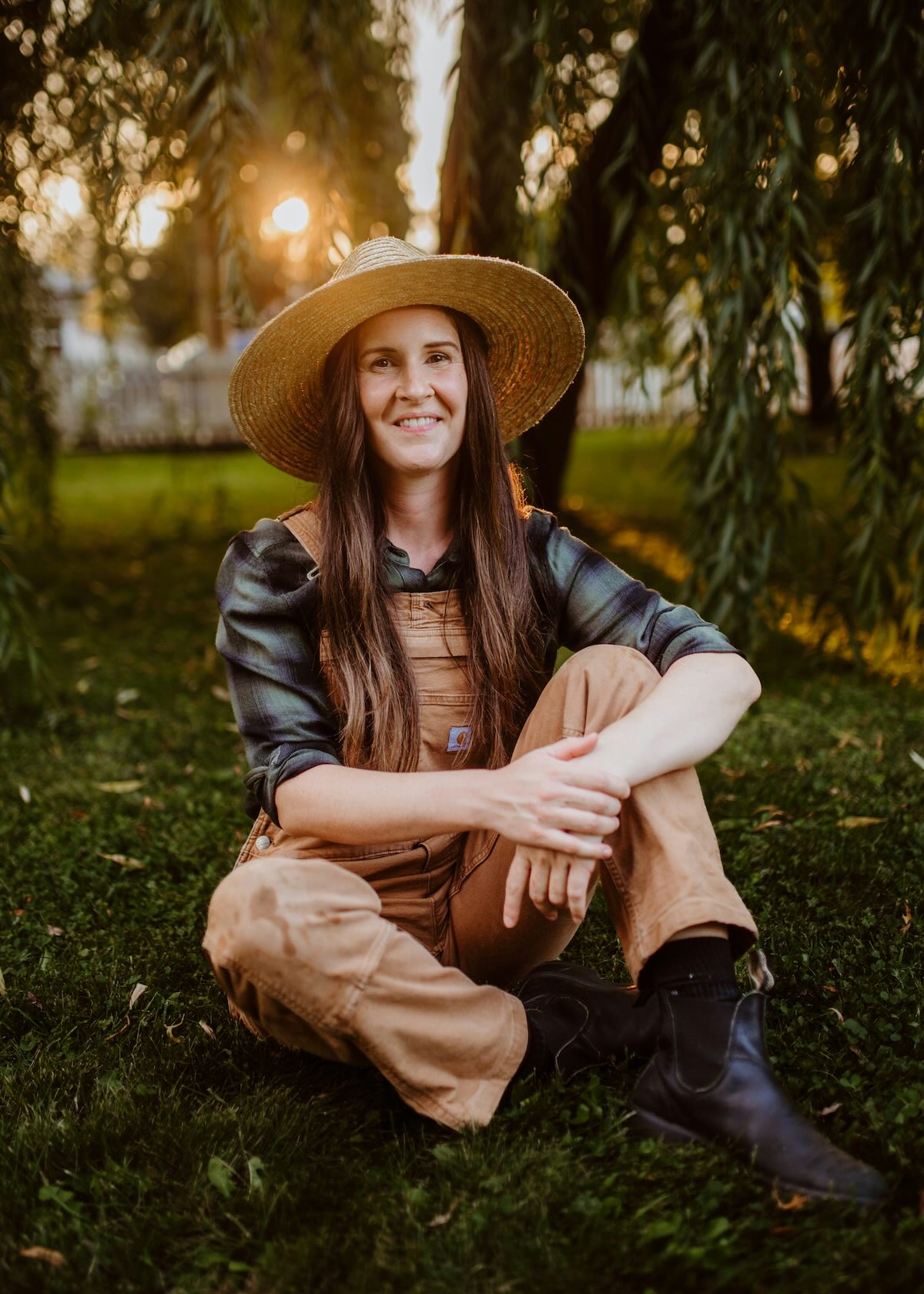 Woman landscaper sitting cross-legged on grass at golden hour, wearing a straw hat, tan overalls, and black boots beneath willow branches, smiling softly.