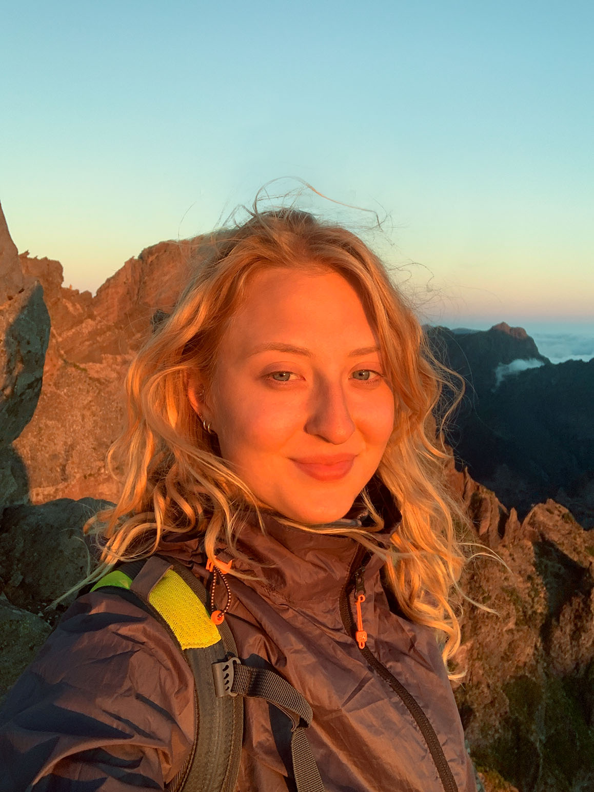 Smiling woman with blonde hair wearing a jacket and backpack, standing outdoors on rocky mountains during sunset.