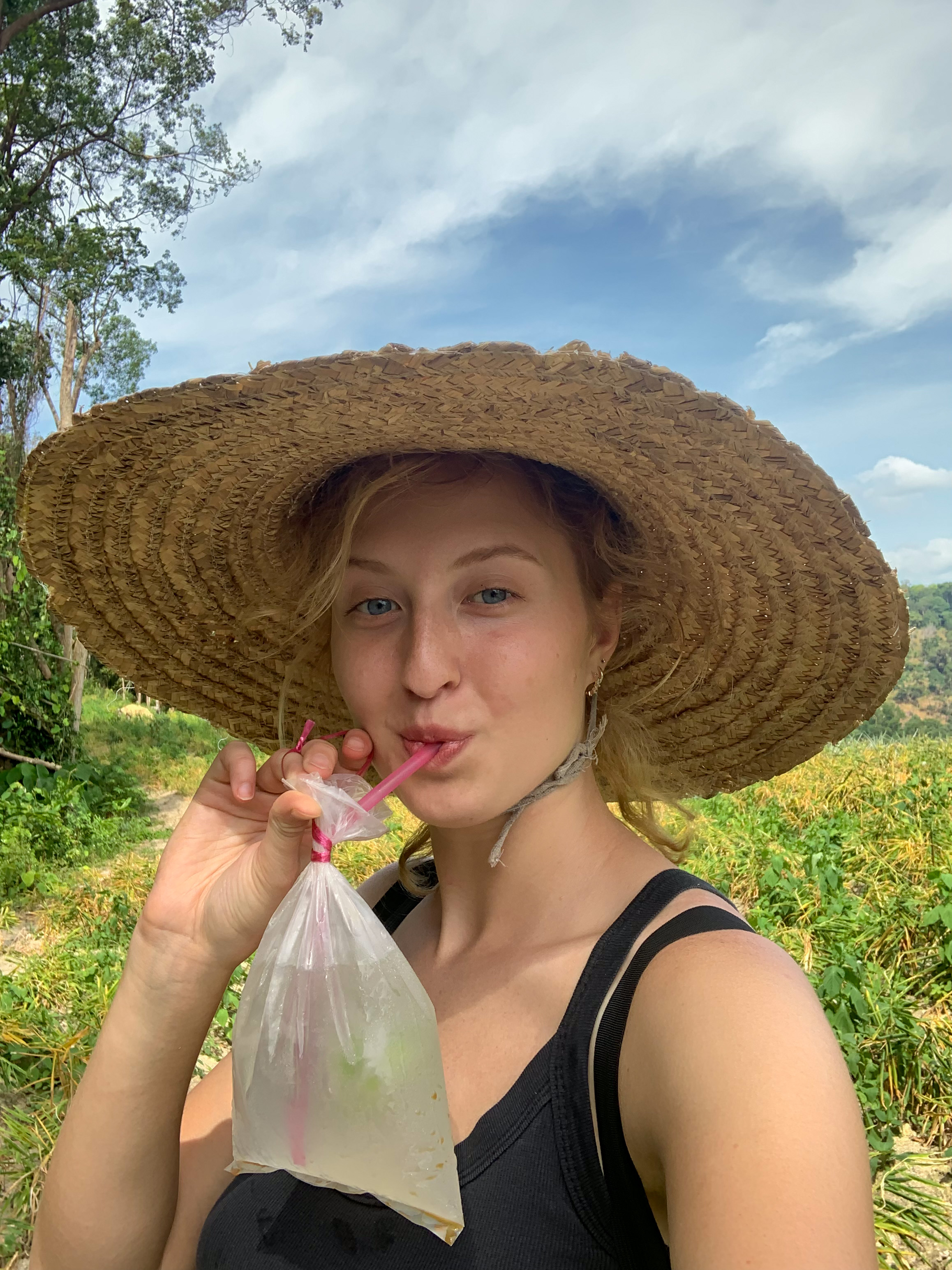 Young woman wearing a large straw hat drinking from a plastic bag with a pink straw in a sunny outdoor green field.