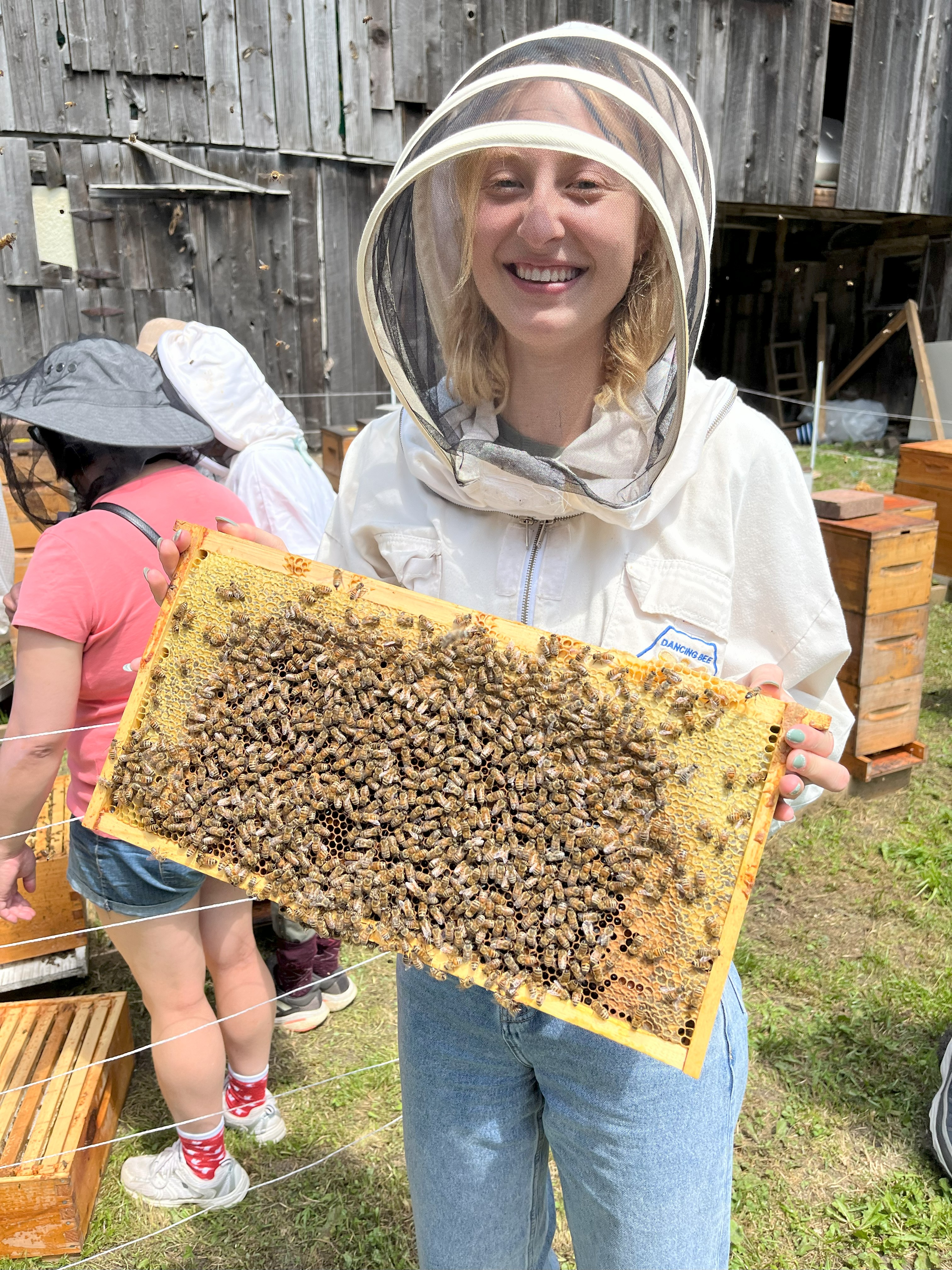 Smiling person in beekeeping suit holding a honeycomb frame covered with bees outdoors.