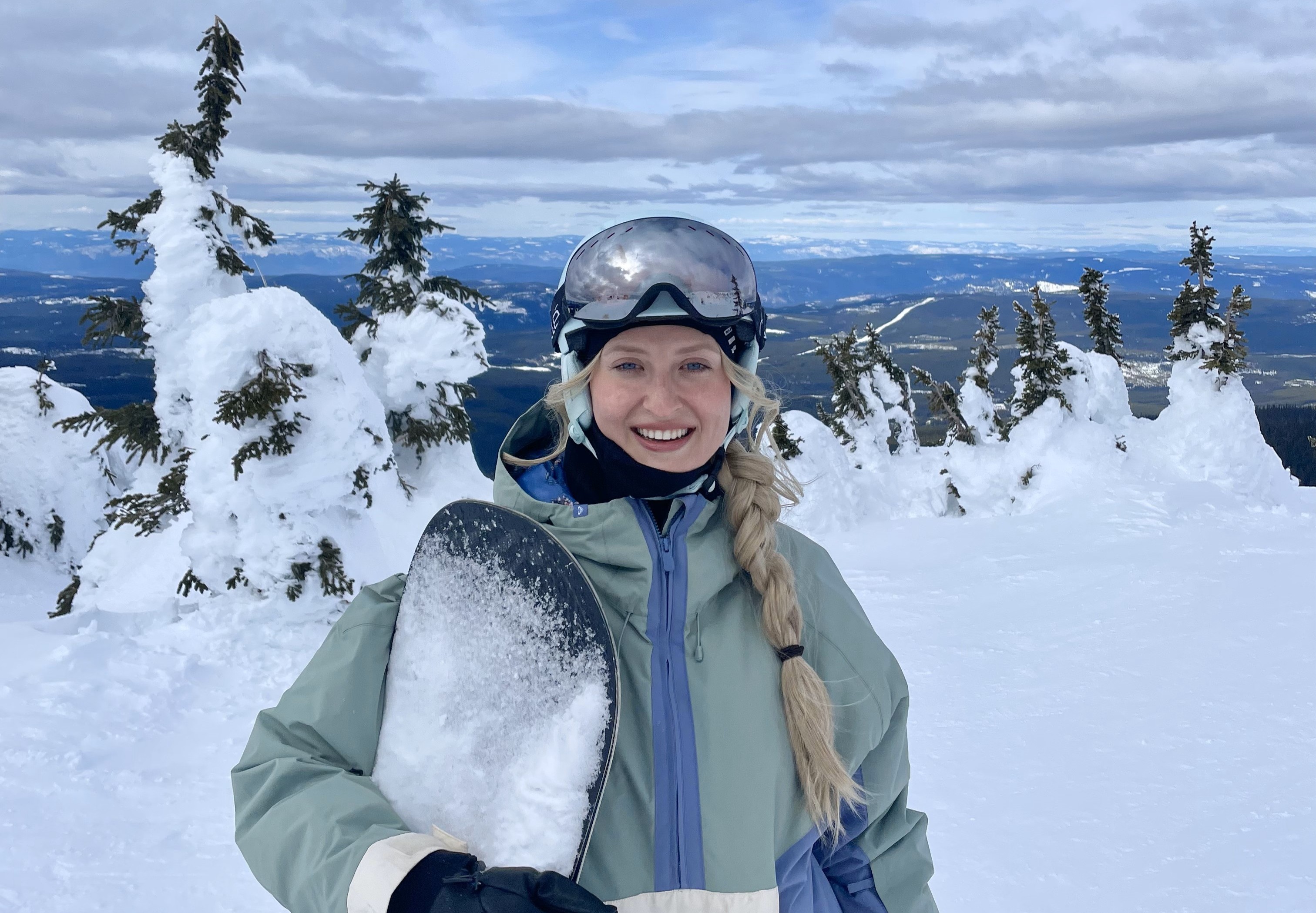 Smiling woman in winter gear holding a snow-covered snowboard on a snowy mountain with snow-laden trees and a distant mountain range in the background.