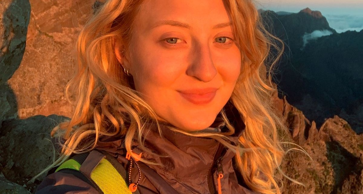 Close-up portrait of a smiling woman with blonde hair illuminated by warm sunlight, with mountain cliffs in the background.