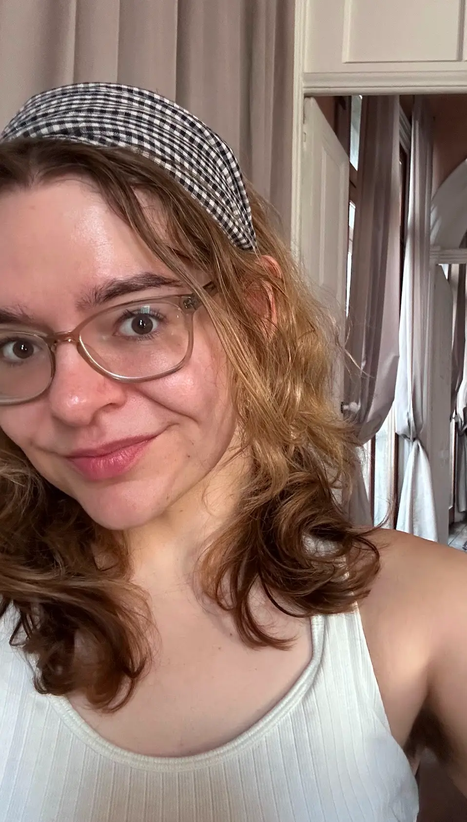 Smiling woman with glasses and a checkered headband, wearing a white ribbed tank top indoors with beige curtains and white doors in the background.