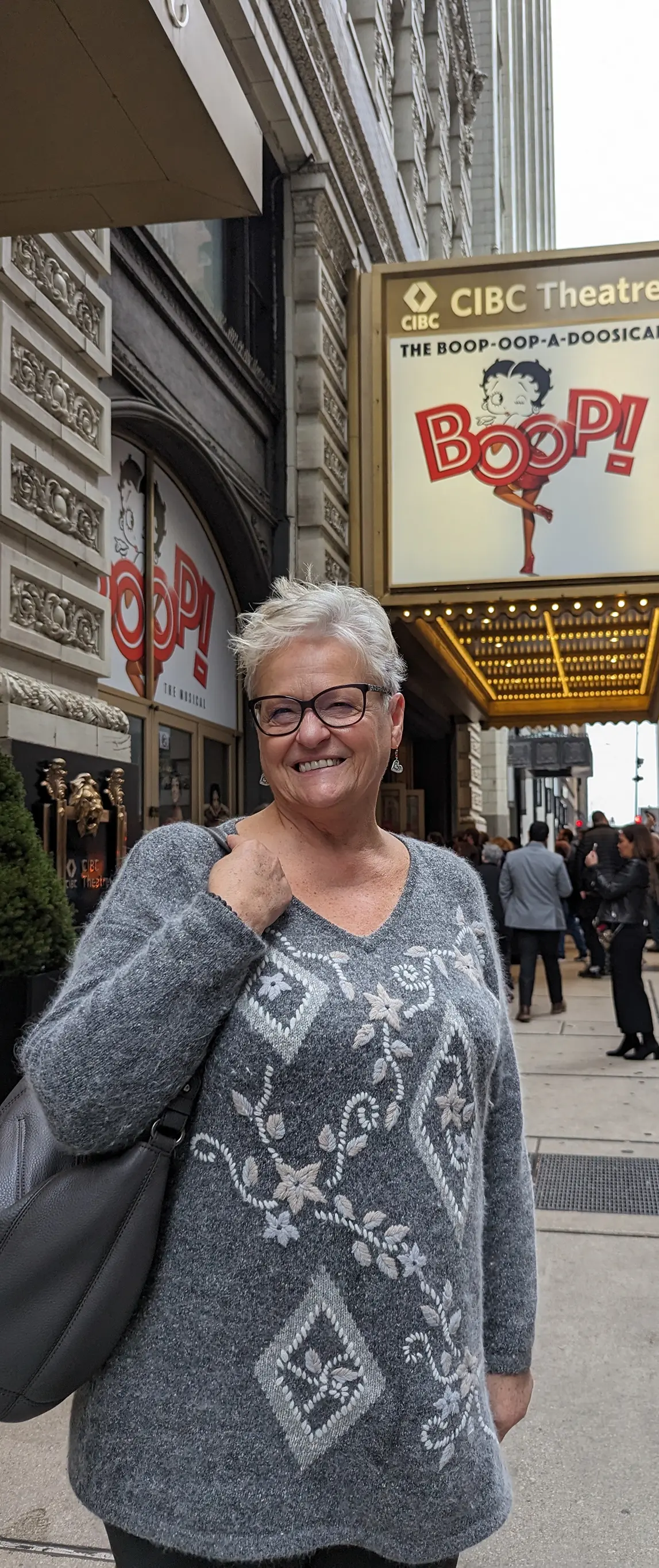 Smiling older woman with short gray hair and glasses wearing a gray floral sweater standing outside the CIBC Theatre with a BOOP! musical sign.