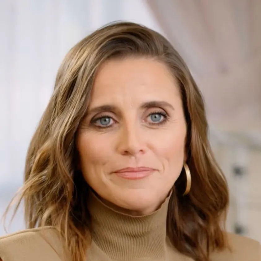 Close-up portrait of a woman with light brown hair wearing a beige turtleneck and gold hoop earrings, smiling softly.