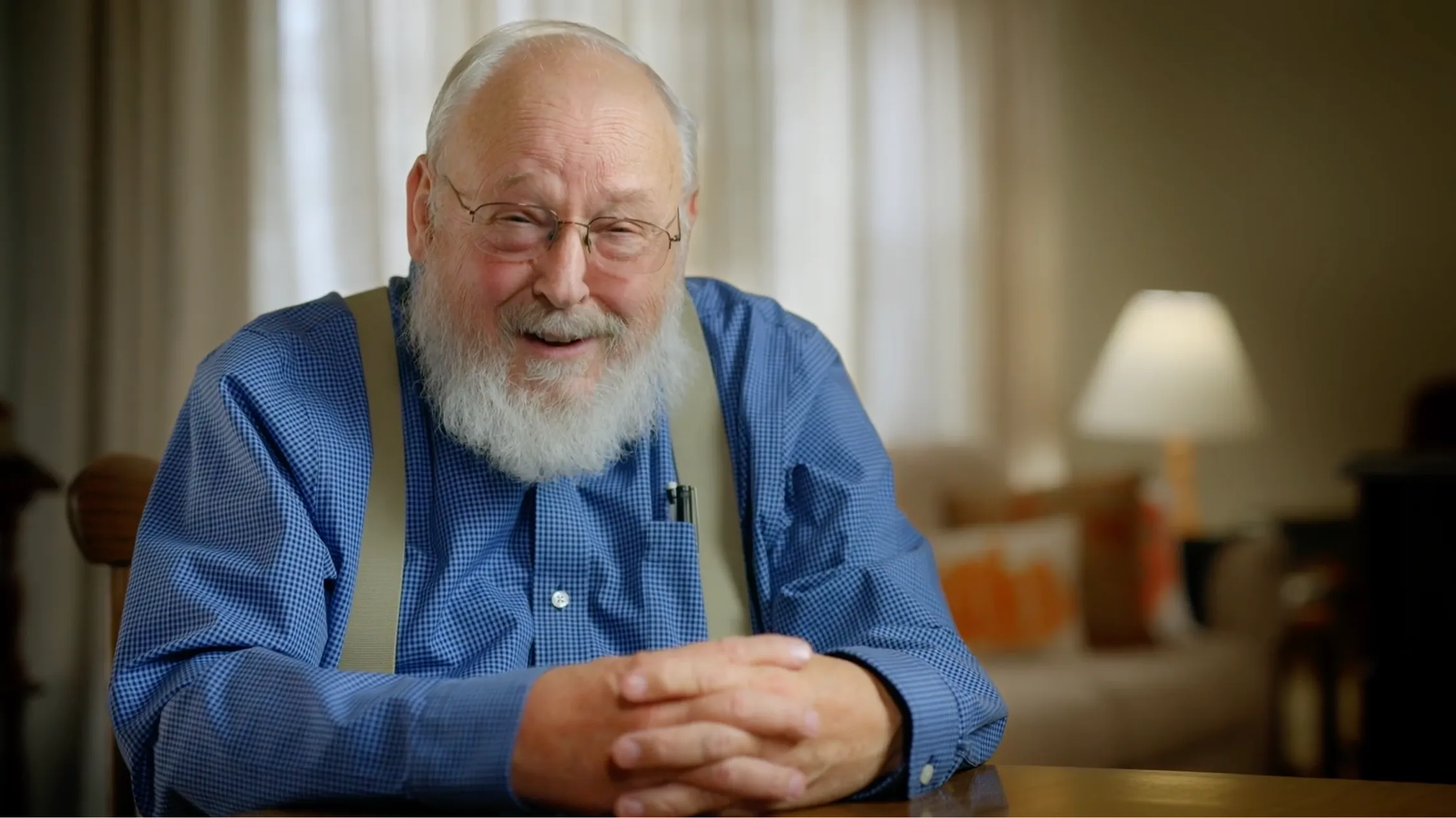 Older man with white beard and glasses wearing a blue checkered shirt and suspenders, smiling while seated at a table.