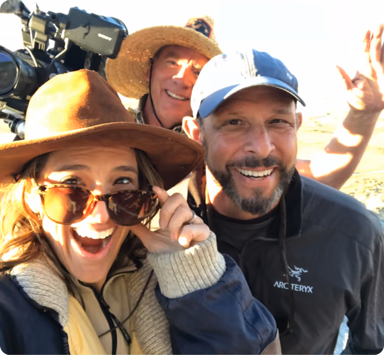 Three smiling people outdoors: a woman in a brown hat adjusting her sunglasses, a man in a gray and blue cap and black jacket with an Arc'teryx logo, and a man behind them wearing a straw hat and waving, with a video camera partially visible.