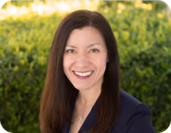 Smiling woman with long brown hair wearing a dark blazer and a necklace, standing outdoors with green foliage in the background.