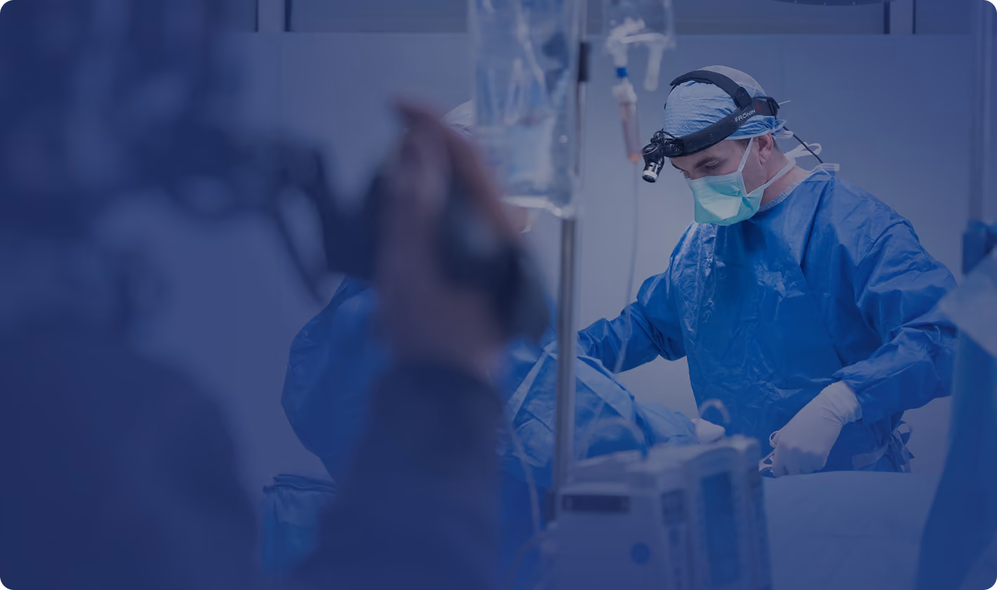 Surgeon in blue surgical gown, mask, and headlight performing an operation in an operating room with medical equipment in the foreground.