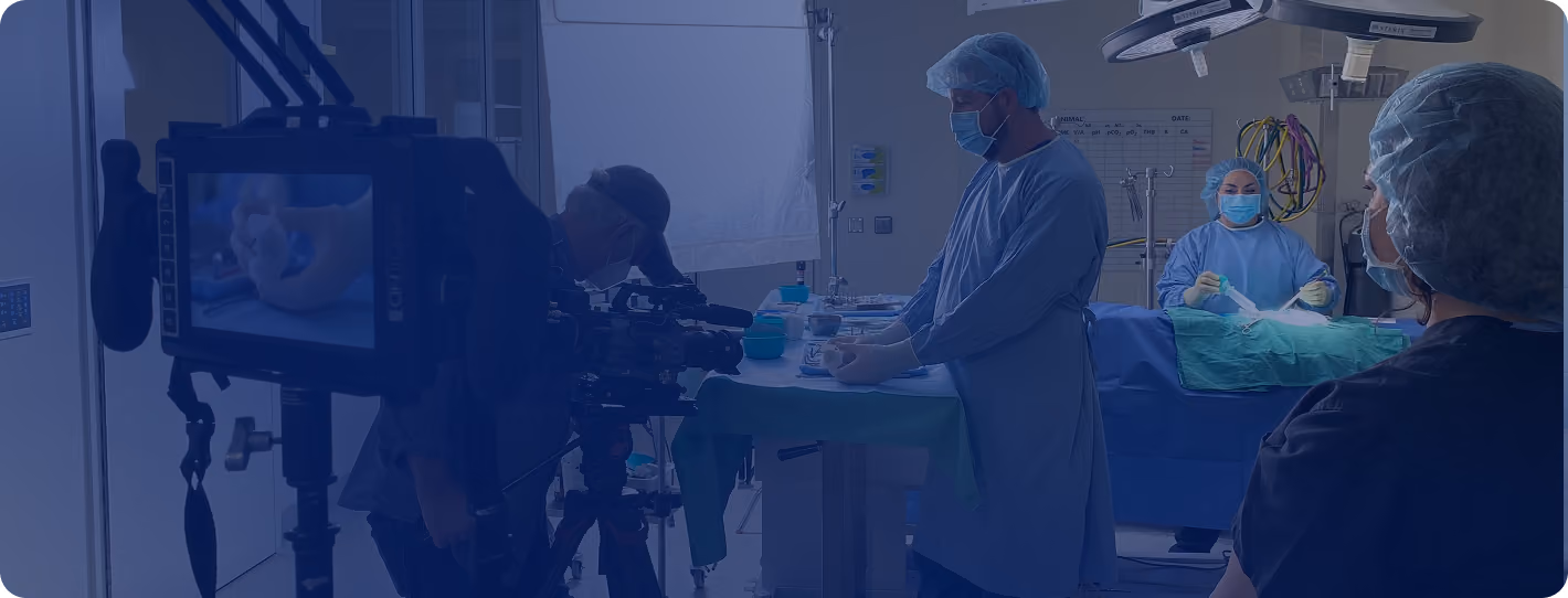 Medical professionals in surgical gowns, masks, and hair covers engaged in a procedure in an operating room with a camera crew filming the activity.