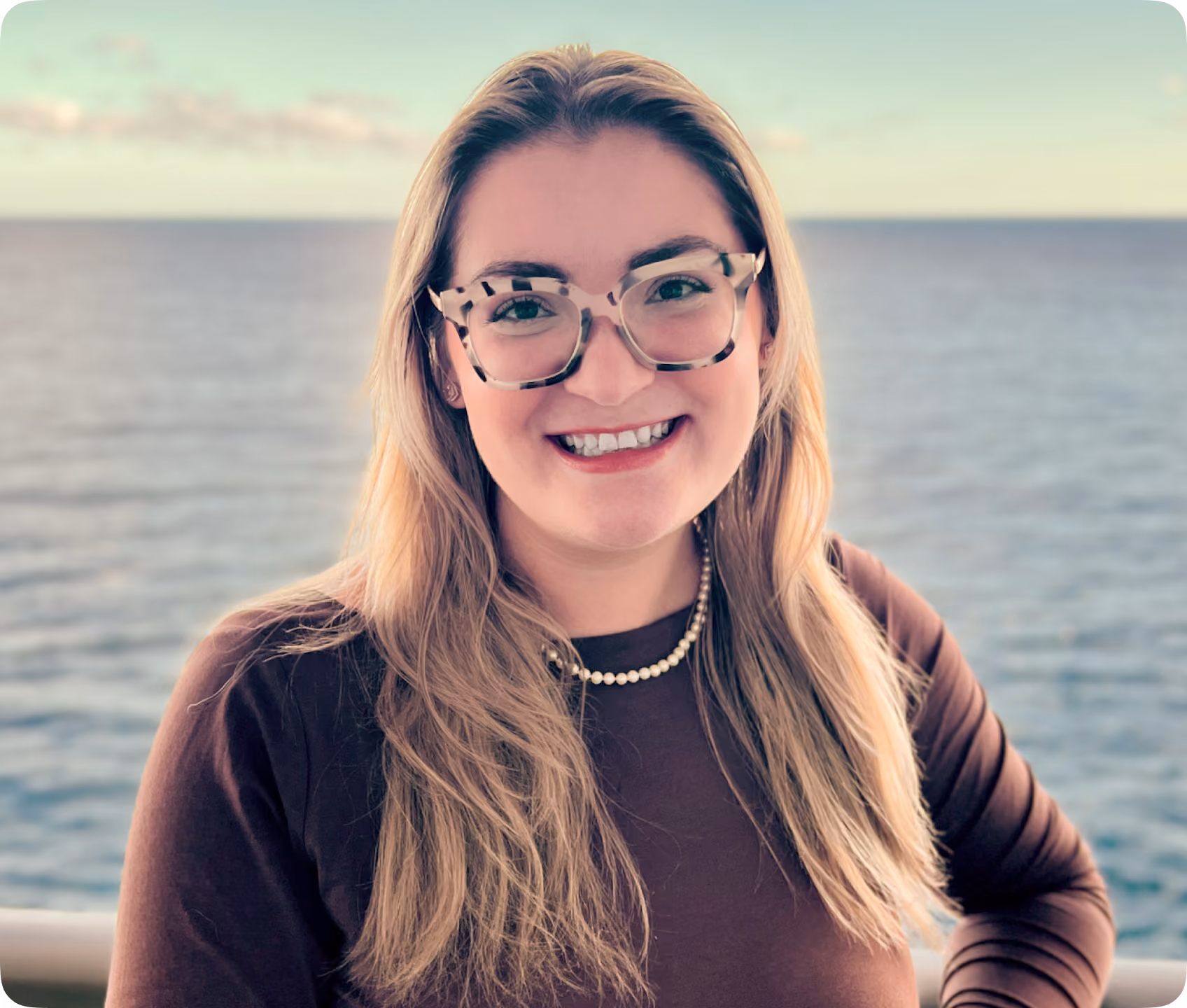 Smiling woman with long blonde hair wearing large tortoiseshell glasses, a brown top, and a pearl necklace standing outdoors with a body of water in the background.