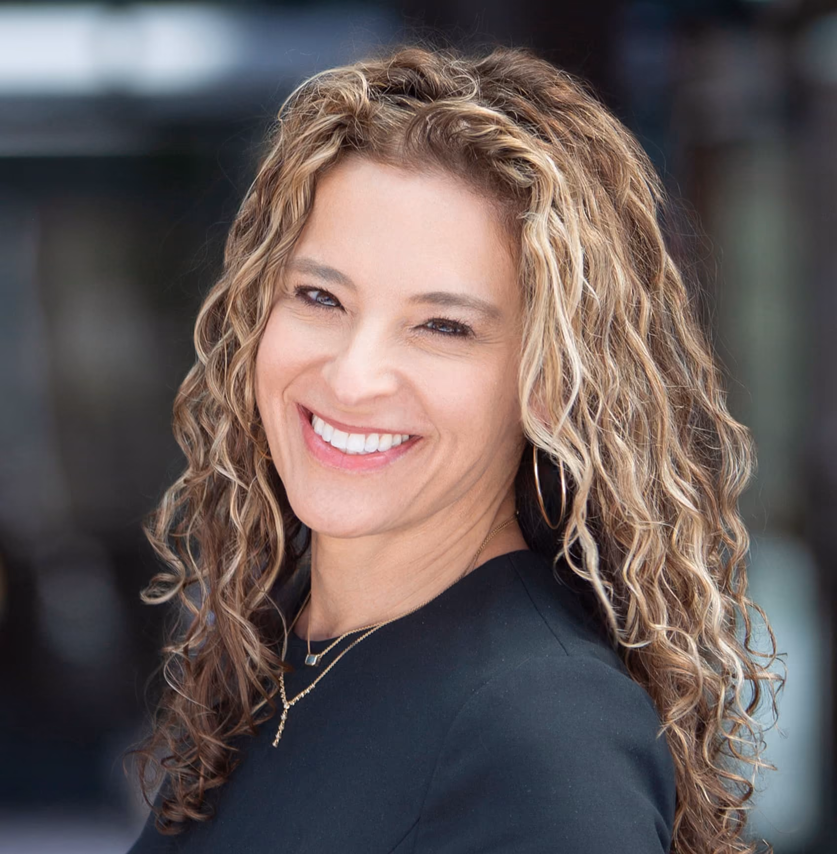 Smiling woman with curly, highlighted hair wearing hoop earrings, a black top, and layered gold necklaces against a blurred background.