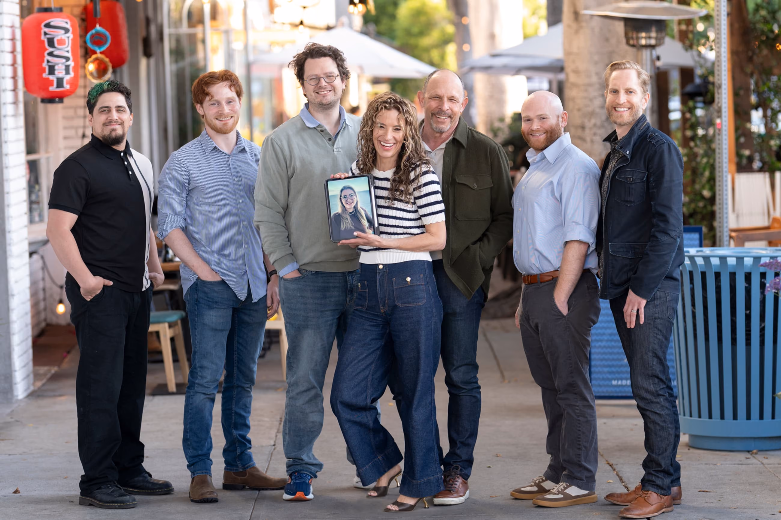 Group of seven people standing on a city sidewalk outside a sushi restaurant, smiling at the camera, with one woman holding a tablet showing an eighth person’s image.