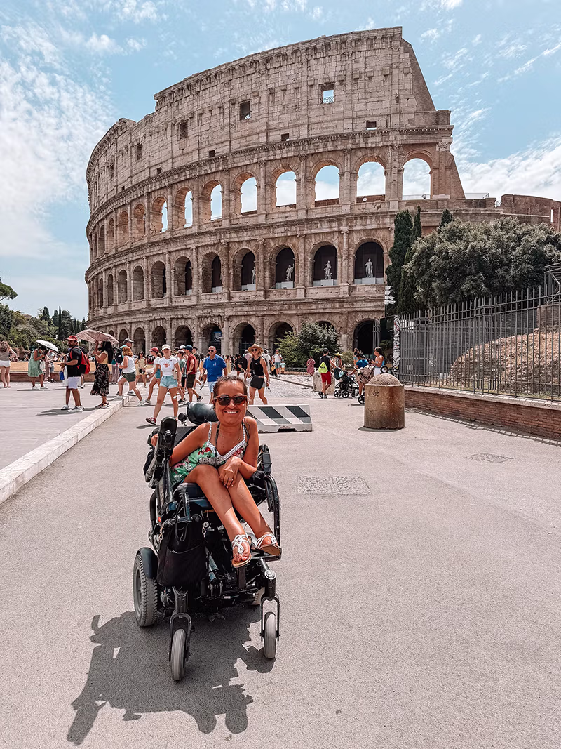 A girl in a wheelchair smiling in front of the Roman Colloseum
