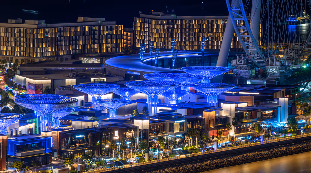 Vibrant night view of Bluewaters Island in Dubai, showcasing a lively dining and entertainment area illuminated with blue lighting, with Ain Dubai observation wheel in the background.