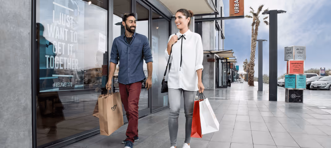 Two men walking outdoors with shopping bags in hand, passing by storefronts with glass windows and palm trees in the background.