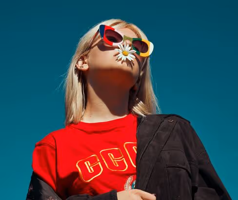 Stylish woman in a red shirt and colorful sunglasses posing confidently against a clear blue sky.
