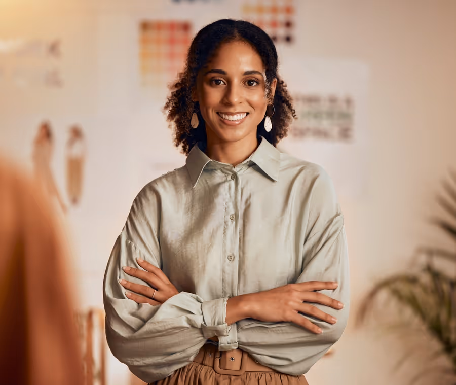 Confident woman smiling with arms crossed, standing in a creative workspace with design boards in the background.