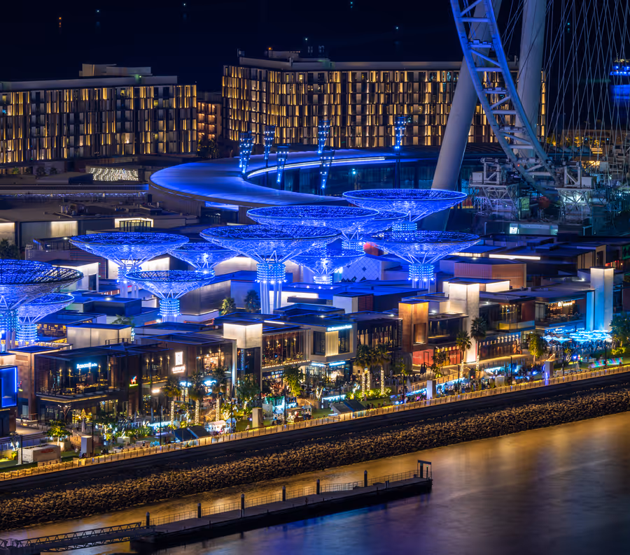 Night view of Bluewaters Island Dubai with illuminated buildings, glowing blue canopies, and Ain Dubai observation wheel.