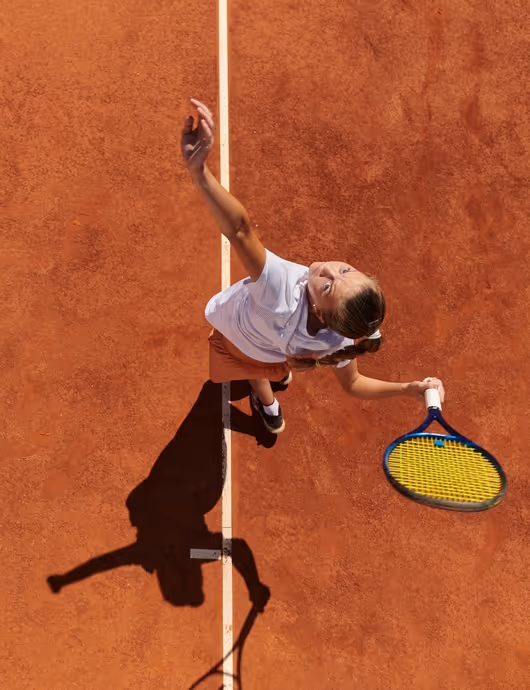Overhead view of a tennis player in mid-serve on a clay court, holding a yellow racket with their shadow visible on the ground.