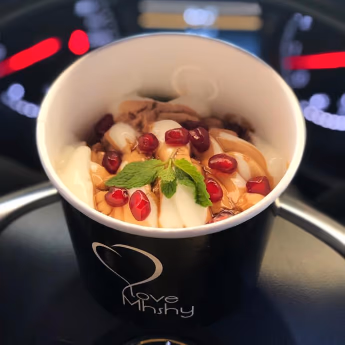 Cup of frozen yogurt topped with pomegranate seeds, syrup, and mint leaves, served in a black branded container.