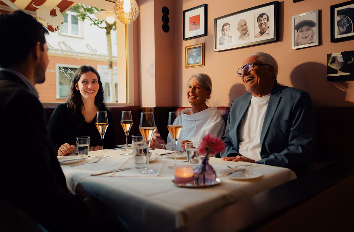 Vier Menschen unterschiedlichen Alters sitzen in einem Restaurant an einem mit weißer Tischdecke gedeckten Tisch und lachen gemeinsam.