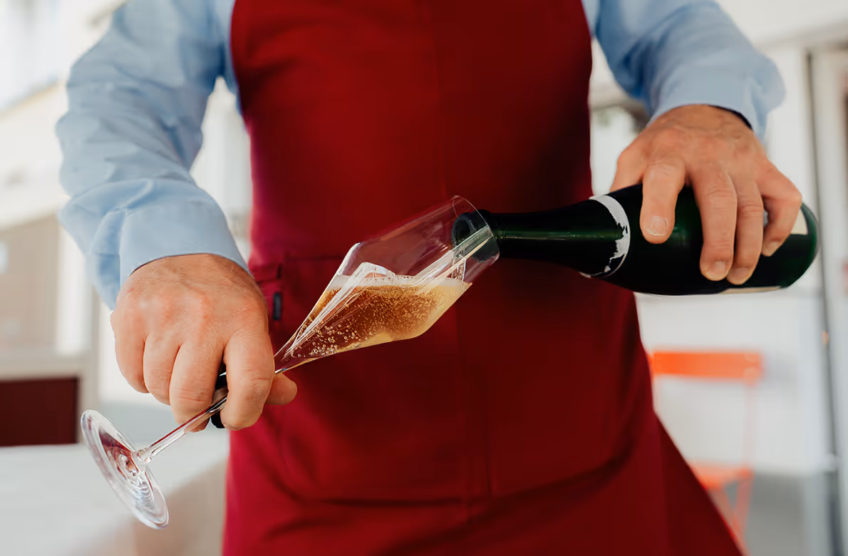 Person in roter Schürze gießt Sekt in ein Glas.