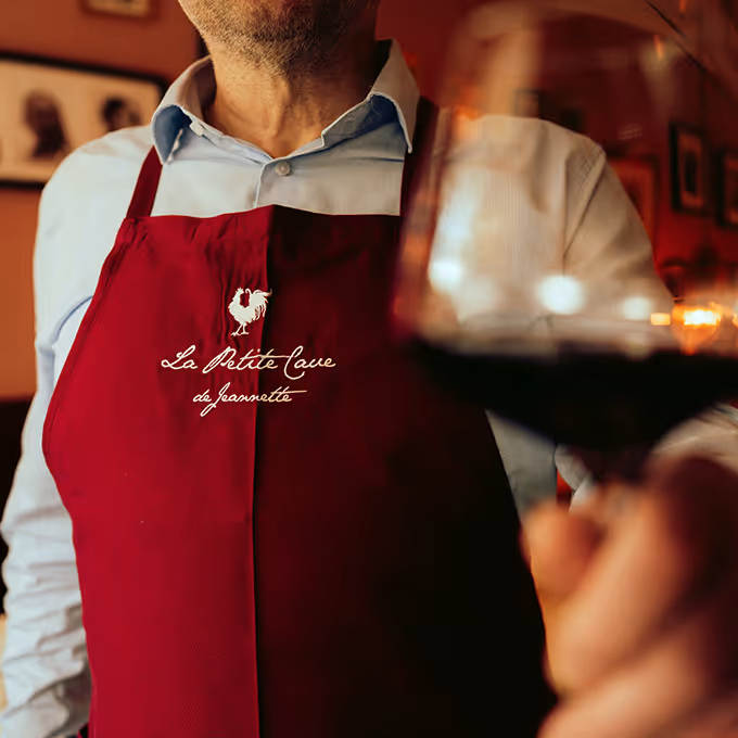 Mann in roter Schürze mit Schriftzug 'La Petite Cave de Jeannette' hält ein Glas Rotwein in einem gemütlichen Restaurant.